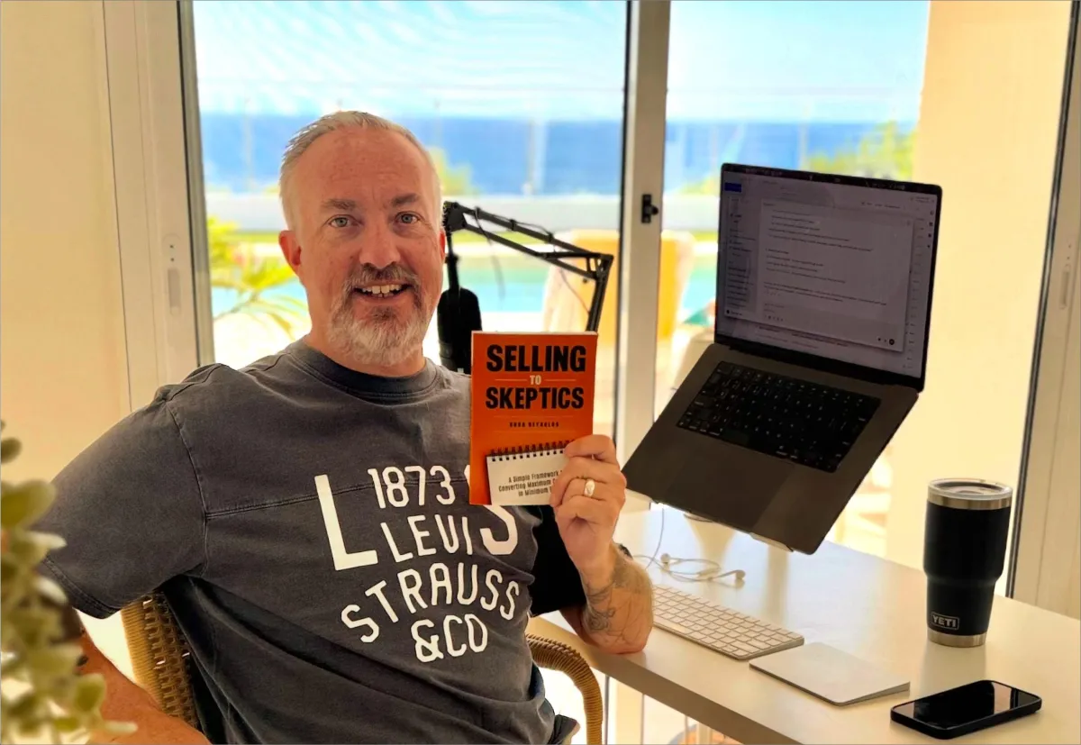 Russ Reynolds at his desk holding a copy of Selling to Skeptics, with the Mediterranean visible through the window behind him.